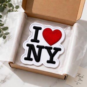 May include: A white embroidered patch featuring the black text I and NY with a red heart symbol in between. The patch has a white border and is placed inside a brown cardboard box lined with white tissue paper, presented for shipping.