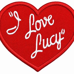 May include: A heart-shaped embroidered patch in bright red with a stitched border. The text "I Love Lucy" is embroidered in white cursive script across the centre of the heart, featuring a textured fabric finish throughout the entire design.