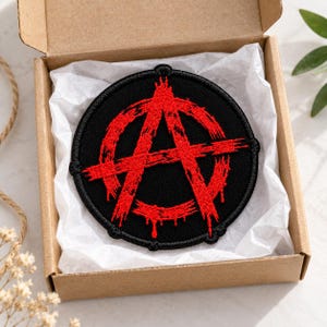 以下が含まれることがあります： 黒い背景に赤いアナーキーシンボルが刺繍された円形のワッペン。デザインはペンキが垂れたようなダメージ加工が施されています。ワッペンは白い薄紙が敷かれた小さな開いた段ボール箱に入っています。