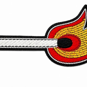 May include: An embroidered patch shaped like a lit matchstick. The matchstick has a white shaft with a black outline, a black head, and a flame depicted in vibrant yellow and red thread. The entire design is bordered by a thick black stitched edge.