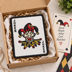 May include: A rectangular embroidered patch featuring a white playing card design with a central jester character in red, black, and yellow. The word JOKER is embroidered in black along the left and right sides. The patch is inside a brown box.