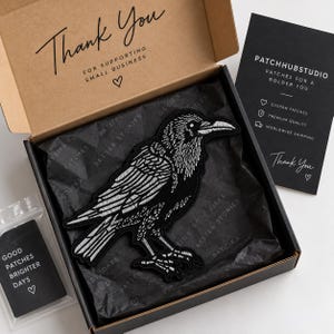 Puede incluir: Un parche bordado negro de un cuervo con detalles de plumas blancas, presentado dentro de una caja de cartón forrada con papel de seda negro. La tapa incluye el texto Thank You For Supporting Small Business y una tarjeta con Patchhubstudio.