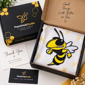 May include: A black cardboard box containing a yellow and black embroidered wasp patch on white tissue paper. The box lid features a bee logo and the text PatchHubStudio. A thank you card and a business card are included with the packaging.