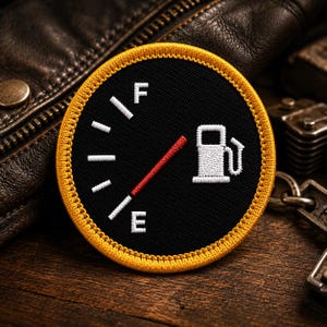 May include: A circular embroidered patch featuring a black background with a white fuel gauge graphic. The design includes the letters F and E, white tick marks, and a red needle pointing between them, all bordered by a thick yellow stitched edge.