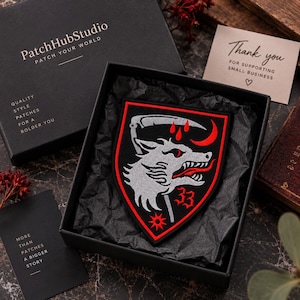 Puede incluir: Un parche bordado en forma de escudo con una cabeza de lobo gris, una guadaña, una luna creciente roja, tres gotas rojas y el número 33 en rojo sobre un fondo negro, presentado dentro de una caja de regalo negra con papel de seda negro.