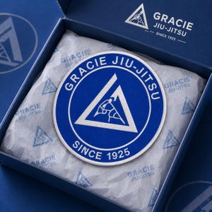 Puede incluir: Un parche circular bordado en azul y blanco con el logotipo de Gracie Jiu-Jitsu y el texto "GRACIE JIU-JITSU SINCE 1925". El parche descansa sobre papel de seda con marca dentro de una caja de cartón azul con el mismo logo en la tapa.