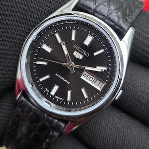 Op de afbeelding: Een Seiko 5 automatisch polshorloge met een ronde zilverkleurige roestvrijstalen kast en een zwarte wijzerplaat. De wijzerplaat is voorzien van zilverkleurige uurmarkeringen, lichtgevende wijzers en een dag-datumvenster op de drie-uurpositie.