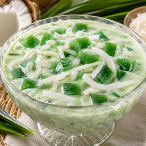 May include: A glass bowl filled with a creamy light green dessert containing translucent dark green gelatin cubes and thin white strips of coconut meat. The mixture has a smooth, liquid consistency and is served in a clear glass dish.