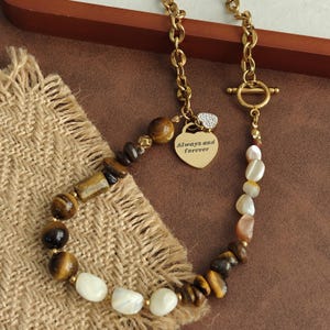 May include: A gold-toned chain necklace featuring a toggle clasp and a heart-shaped pendant engraved with the text Always and forever. The necklace is adorned with a mix of polished tiger eye beads and smooth white shell-like stones in various shapes.