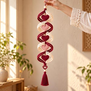 May include: A vertical hanging decoration featuring a spiral design made of alternating cream and deep red crocheted ribbons. Five crocheted hearts in shades of red and light pink are centered within the spiral. It ends with a red tassel.