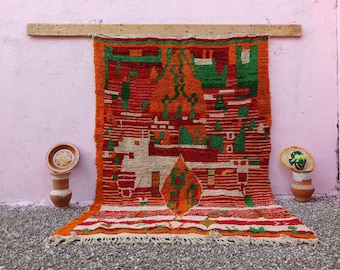 May include: A rectangular hand-woven rug featuring a vibrant color palette of orange, red, and green with a central white animal figure. The rug displays geometric patterns, horizontal stripes, and a fringed edge at the bottom, hanging from a wooden beam.