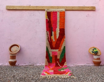 May include: A long, rectangular runner rug featuring a vibrant, multi-colored geometric pattern in shades of hot pink, deep red, bright orange, green, and cream. The rug has a textured, knotted pile and short fringe at the bottom edge.