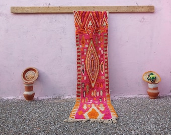 May include: A long, narrow runner rug featuring a vibrant fuchsia pink base with intricate geometric patterns in orange, yellow, and white. The design includes a large central diamond motif, smaller scattered shapes, and a fringed edge at the bottom.