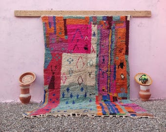 May include: A rectangular area rug featuring a vibrant, multi-colored geometric pattern with blocks of hot pink, orange, blue, and cream. The rug displays various small, dark-colored diamond and abstract symbols throughout, finished with a fringed edge.