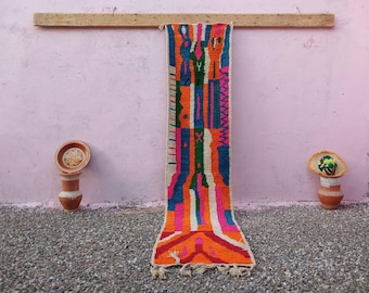 May include: A long, narrow, hand-woven runner rug featuring a vibrant, multi-colored geometric pattern in shades of bright orange, hot pink, deep blue, green, and cream. The rug has a fringed bottom edge and is displayed hanging vertically.