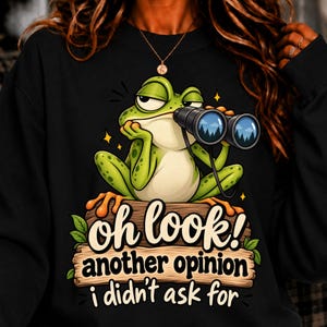 May include: A black long-sleeve sweatshirt featuring a graphic of a green frog holding black binoculars while sitting on a wooden log. The text reads, "oh look! another opinion i didn't ask for" in a stylized font with small green leaves.