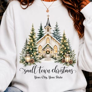 Może przedstawiać: Biała bluza z okrągłym dekoltem z grafiką kościoła otoczonego ośnieżonymi sosnami, latarniami i szyszkami. Napis "small town christmas" wykonano kursywą, a poniżej znajduje się tekst "Your City, Your State" czcionką szeryfową.