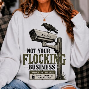 May include: A white long-sleeve sweatshirt featuring a graphic of a black crow perched on a security camera. The text reads "NOT YOUR FLOCKING BUSINESS" and "PRIVACY ISN'T PARANOIA I DON'T CONSENT TO BEING WATCHED" with a small padlock icon.