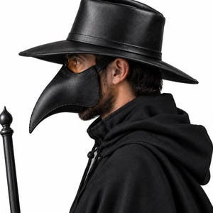 Puede incluir: Una máscara de médico de la peste negra con un pico largo y puntiagudo y un sombrero negro de ala ancha a juego. La máscara cuenta con lentes oscuros y se lleva con una capa negra con capucha. Un bastón negro con remate redondo es visible.