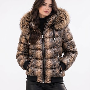 Könnte beinhalten: Eine Steppjacke mit braun-schwarzem Schlangenmuster, durchgehendem Frontreißverschluss und einer Kapuze mit dickem, braunem Kunstfellbesatz. Die Jacke verfügt über schwarze Rippstrickbündchen an den Ärmeln und am Saum sowie zwei Reißverschlusstaschen.