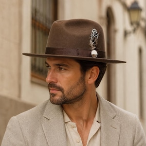 Puede incluir: Un sombrero fedora de fieltro marrón con una cinta de grosgrain marrón oscuro, un broche circular plateado y una pluma moteada en blanco y negro insertada en el lateral de la copa. El sombrero tiene un ala estructurada y una copa pellizcada.