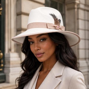 May include: A white fedora hat featuring a wide beige ribbon band with a small bow detail. The hat is accented with a speckled brown and white feather tucked into the side of the band, which also includes a small circular pink decorative pin.