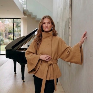 Può includere: Una giacca color cammello con collo alto, ampie maniche a campana e una cintura in tessuto abbinata in vita. Il capo presenta una silhouette strutturata ed è abbinato a semplici pantaloni neri per un look pulito ed essenziale.