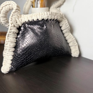 Könnte beinhalten: Eine Handtasche mit einem schwarzen Körper aus Kunst-Schlangenleder mit texturiertem Schuppenmuster. Die Tasche ist mit einem dicken, cremefarbenen gehäkelten Kordelbesatz eingefasst, der in zwei abgerundete Griffe an den Oberkanten übergeht.