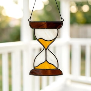 May include: A stained glass suncatcher shaped like an hourglass, featuring dark brown top and bottom sections, white glass panels, and vibrant amber-colored glass representing sand, all held together by dark metal cames and a black hanging cord.