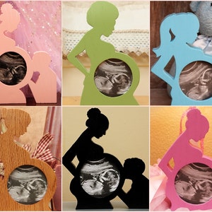 May include: Six wooden picture frames shaped like a pregnant woman's silhouette. Each frame is a different color: pink, green, blue, brown, black, and light pink. The frames have a circular opening in the center to hold an ultrasound image.