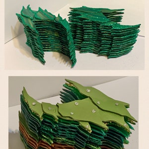 May include: A stack of green, gold, and copper glitter paper scales. The scales are layered and have a slightly curved shape. The scales are arranged in a pattern that resembles a dragon's tail.