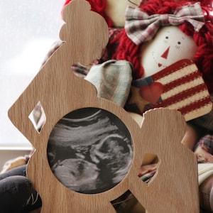 May include: Wooden cutout of a pregnant person holding a sonogram photo in a circular frame. The cutout is light brown and has a rustic appearance.