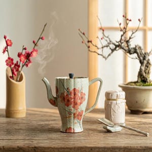 Peut inclure: Une théière en céramique de couleur claire avec un corps cannelé et un motif floral rouge peint. La théière présente un bec verseur incurvé, une anse et un petit bouton de couvercle sombre. De la vapeur s'échappe et une cuillère repose.