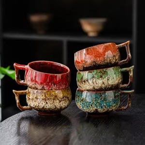 May include: A set of five ceramic mugs with a reactive glaze finish, arranged in two stacks. The mugs feature a gradient colour effect, transitioning from deep, saturated hues at the top to a textured, speckled cream base with angular handles.