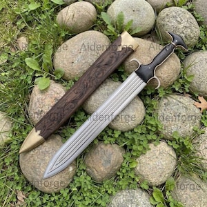 May include: A double-edged dagger with a fluted steel blade and a dark wooden hilt featuring a curved pommel. The matching wooden scabbard is adorned with intricate scrollwork engravings and finished with a polished brass tip and throat.