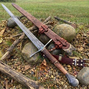 Può includere: Una spada in acciaio a doppio taglio con una lama finemente incisa, una guardia con una piccola pietra verde e un'impugnatura rivestita in pelle marrone. Include un fodero in pelle marrone abbinato con motivi in rilievo e una cintura.