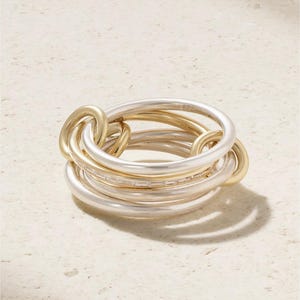 Op de afbeelding: Een ring met meerdere banden bestaande uit in elkaar grijpende zilver- en goudkleurige cirkelvormige banden. Een van de zilveren banden is ingelegd met een rij kleine, heldere baguette-geslepen stenen die het licht vangen voor een gelaagd effect.