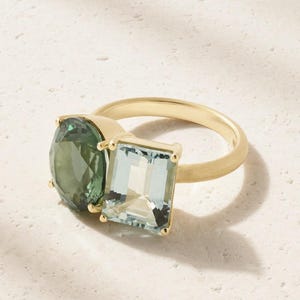 May include: A gold ring featuring two prominent gemstones set side by side. One stone is a round, deep green gem, while the other is a rectangular, light blue-green gem. Both stones are held in place by gold prongs on a smooth gold band.