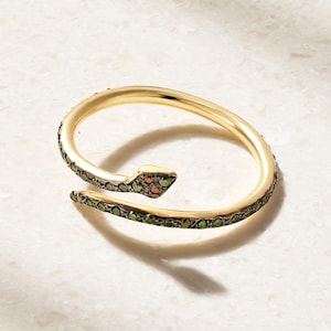May include: A gold-toned snake-shaped ring featuring a wrap-around design. The body of the snake is encrusted with small green stones along the band, while the head is detailed with two small orange stones for the eyes on a dark metal setting.