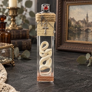 Könnte beinhalten: Eine hohe, rechteckige Glasflasche mit einer gewundenen weißen Schlangenfigur im Inneren. Der Flaschenhals ist mit beigefarbenem Garn umwickelt und mit einem silbernen Ankh-Anhänger sowie kleinen Kreuzen an einer Kette verziert. Der schwarze Verschluss zeigt ein rotes Auge.