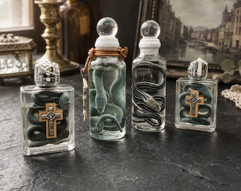 Pode incluir: Quatro garrafas de vidro transparente contendo figuras de cobras enroladas. Duas garrafas apresentam uma cruz de madeira com um crucifixo de prata fixado na frente. Uma garrafa tem um cordão de couro em volta do gargalo com um pingente.