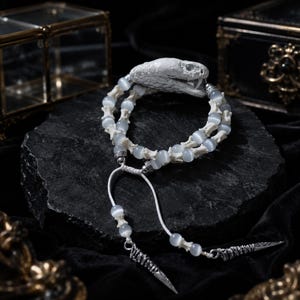 Puede incluir: Una pulsera con una pieza central en forma de cabeza de serpiente blanca, cuentas de ojo de gato gris claro translúcidas y pequeños espaciadores con forma de hueso. Incluye detalles en metal plateado y dos cordones con colgantes puntiagudos.