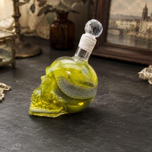 Könnte beinhalten: Eine durchsichtige, totenkopfförmige Glasflasche, gefüllt mit einer leuchtend gelben Flüssigkeit und einer darin aufgerollten grauen Schlange. Der Flaschenhals ist mit weißem Material umwickelt und mit einem facettierten, rautenförmigen Glasstopfen versehen.