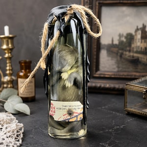 May include: A clear glass bottle containing a small, grey, furry creature figure. The bottle neck is sealed with black wax and wrapped in tan twine. A rectangular paper label is attached to the front with the text, Take this ticket with you during the journey.