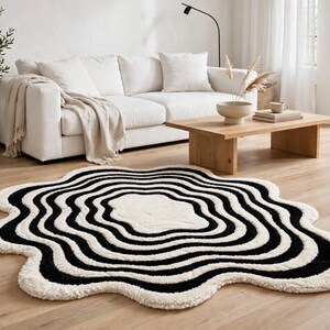 Black and ivory hand-tufted area rug featuring multiple irregular concentric waves surrounding a soft ivory center. The organic-shaped rug has a sculptural scalloped silhouette and plush textured surface. It is displayed in a bright modern living room with a white sofa, natural wood coffee table, indoor olive tree, neutral décor, sheer curtains and warm hardwood flooring.