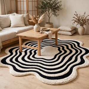 Artistic hand-tufted wavy area rug in black and ivory, designed with flowing concentric organic lines and an irregular scalloped border. The contemporary monochrome rug creates a bold focal point in a minimalist living room featuring a white sectional sofa, natural wood coffee table, indoor olive tree, neutral décor and warm hardwood flooring.