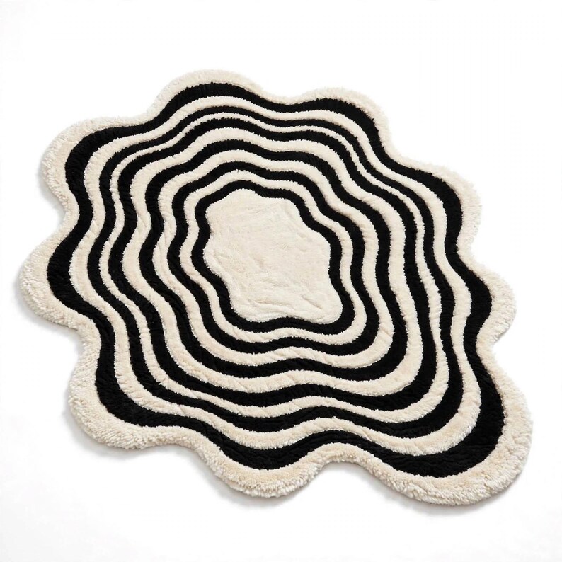 Contemporary black and ivory abstract area rug with a hand-tufted plush texture and sculptural organic shape. The design features flowing irregular black bands surrounding a soft ivory center, creating a ripple-inspired pattern. Shown in a calm neutral living room with white seating, natural wood furniture, greenery, woven accessories and warm hardwood floors.