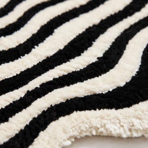 Modern hand-tufted black and white wavy rug with an organic irregular silhouette and layered concentric wave pattern. Alternating black and ivory lines flow around the center, creating a bold optical effect. The statement rug is styled in a minimalist living room with a cream sofa, light oak coffee table, neutral textiles, indoor plants and natural wood flooring.