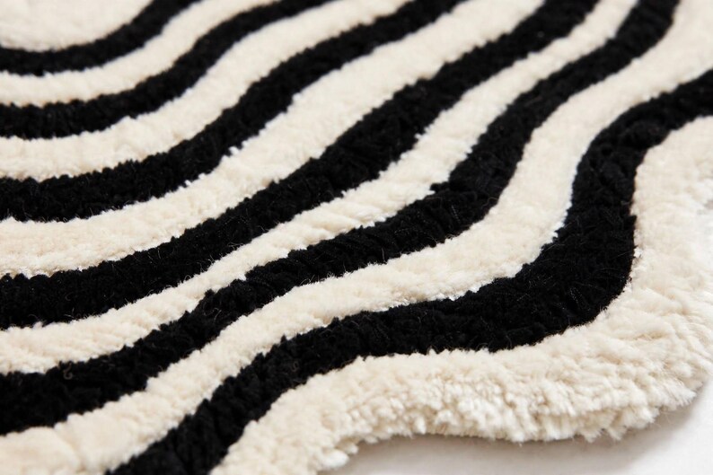 Modern hand-tufted black and white wavy rug with an organic irregular silhouette and layered concentric wave pattern. Alternating black and ivory lines flow around the center, creating a bold optical effect. The statement rug is styled in a minimalist living room with a cream sofa, light oak coffee table, neutral textiles, indoor plants and natural wood flooring.