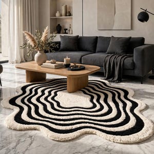 Black and white hand-tufted statement rug with an abstract ripple pattern and irregular sculptural silhouette. The plush rug combines raised ivory and black wave bands around an organic central shape. It complements a modern Scandinavian-inspired interior with a white sofa, light wood furniture, neutral accessories, indoor greenery and warm wooden floors.